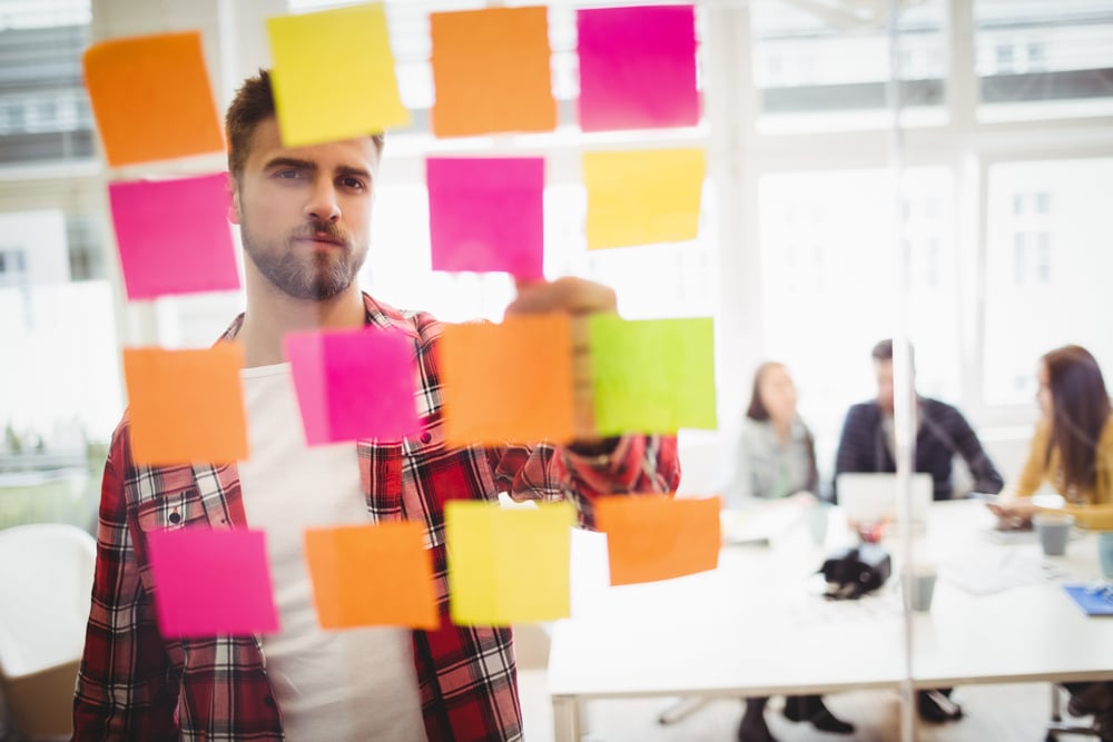 Photo editor looking at multi colored sticky notes on glass in meeting room at creative office